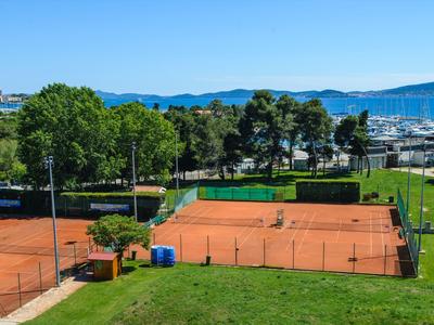 Terrains de tennis en terre battue avec pelouse verte, arbres et port en arrière-plan par temps ensoleillé.
