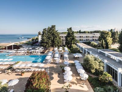 Hotelanlage mit Pool, weißen Sonnenschirmen, Bäumen und Meer im Hintergrund bei klarem Himmel.