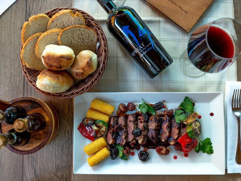 Nakryty stół z czerwonym winem, chlebem, marynowanymi oliwkami i talerzem z grillowanym mięsem i warzywami.