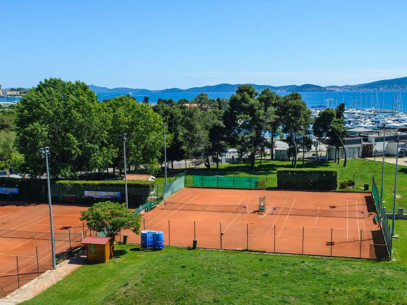 Dwa czerwone korty tenisowe otoczone zielenią z widokiem na marinę w tle.