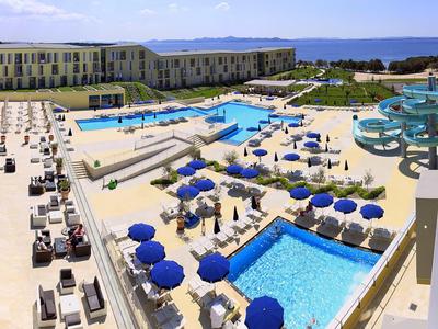Area piscina moderna dell'hotel con più piscine, ombrelloni blu e vista sul mare adiacente.
