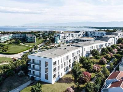 Plusieurs complexes de vacances modernes au bord de la mer avec espaces verts et habitations sous un ciel nuageux.