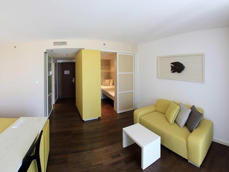 Moderní hotelový pokoj s žlutou pohovkou, světlým stolem a otevřenou skříní.