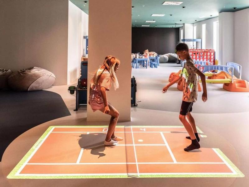 Děti si hrají s interaktivní projekcí na podlaze v moderním vnitřním prostoru.
