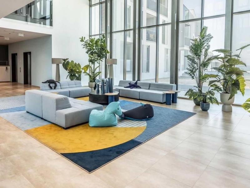 Světlá lounge s pohovkou, kobercem a velkými rostlinami u skleněných oken v hotelu.