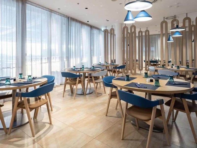 Světla moderní restaurace s modrými židlemi a dřevěnými stoly u velkých oken.