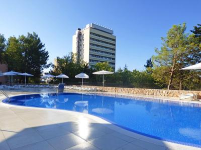 Modern hotel complex with large pool, sun umbrellas, and surrounding trees under clear sky.