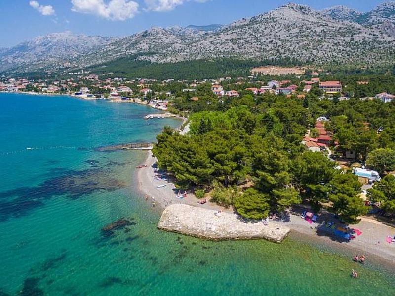 View of a forested coastline with clear blue sea and mountains in the background.