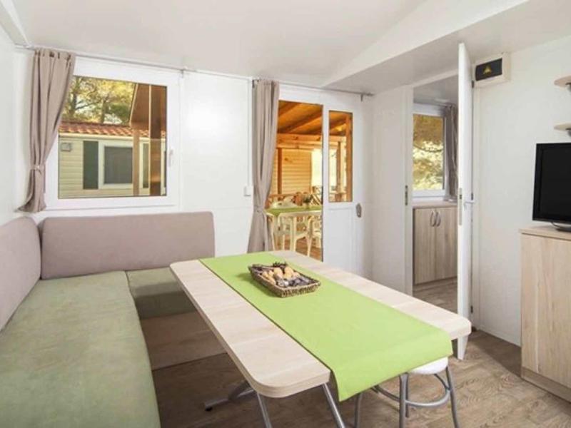 Cozy dining area with corner bench, chairs, green table runner, and TV in a bright room.