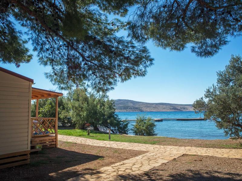 Mobile home with porch under trees by a pebble beach with sea view under clear sky.