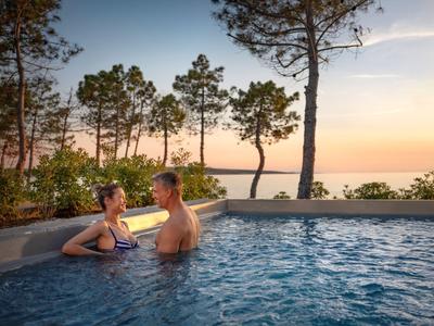 Couple relaxes in an outdoor pool with sunset and trees in the background.