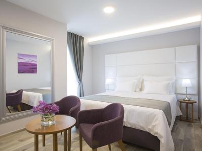 Modern hotel room with large bed, purple armchairs, small table, and wall mirror.