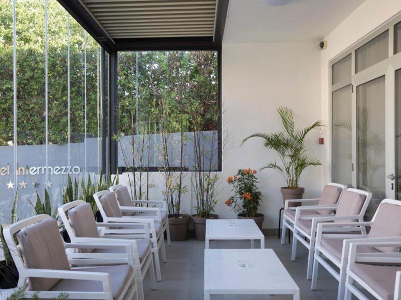 Covered terrace with white armchairs, tables, and plants beside glass wall.