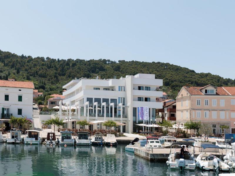 Complexe hôtelier blanc moderne près du port avec des bateaux et une colline boisée en arrière-plan.