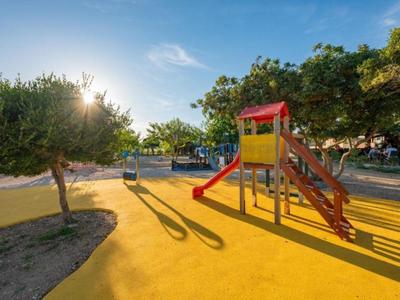 Spielplatz mit Rutsche und Leitern auf gelbem Boden, umgeben von Bäumen und blauen Himmel.
