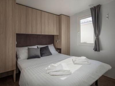 Kleines Schlafzimmer mit Doppelbett, weißen Wänden, Holzmöbeln und einem Fenster mit grauem Vorhang.