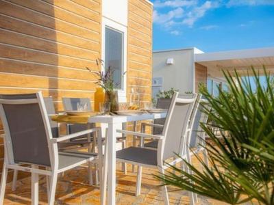 Terrasse mit weißen Stühlen und Tisch neben Holzwand, blauer Himmel im Hintergrund.