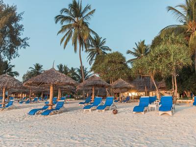 Strand mit weißen Sand, blauen Liegestühlen, Strohdächern und hohen Palmen bei klarem Himmel.