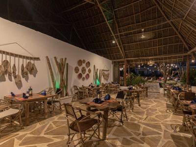 Offenes Restaurant mit Holztischen, Stühlen und Dekoration an weißen Wänden unter Strohdach.