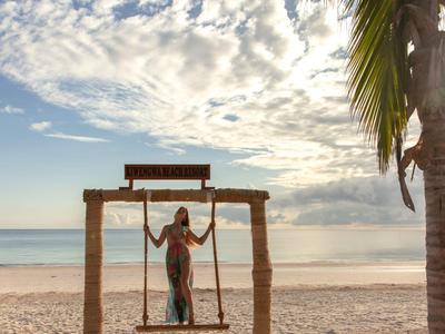 Frau auf Schaukel am Strand, blauer Himmel mit Wolken und Palmenblätter im Vordergrund.