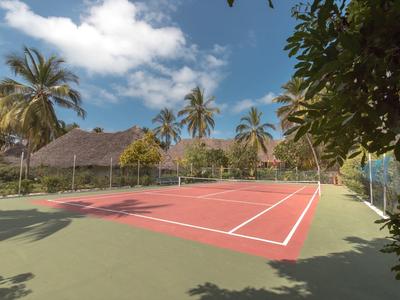 Freiluft-Tennisplatz mit rotem Spielfeld, umgeben von Palmen und Hütten unter blauem Himmel.