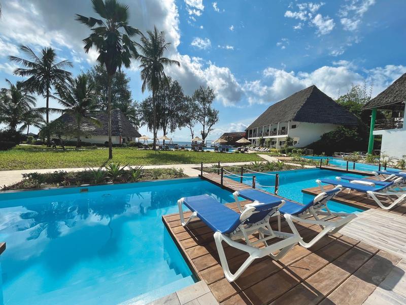 Poolbereich mit Liegestühlen neben einem Hotel unter blauem Himmel und Palmen.