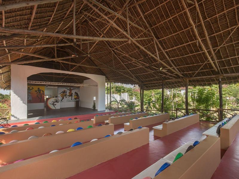 Offener Veranstaltungsraum mit farbigen Bänken und Bühne unter einem Strohdach in tropischer Umgebung