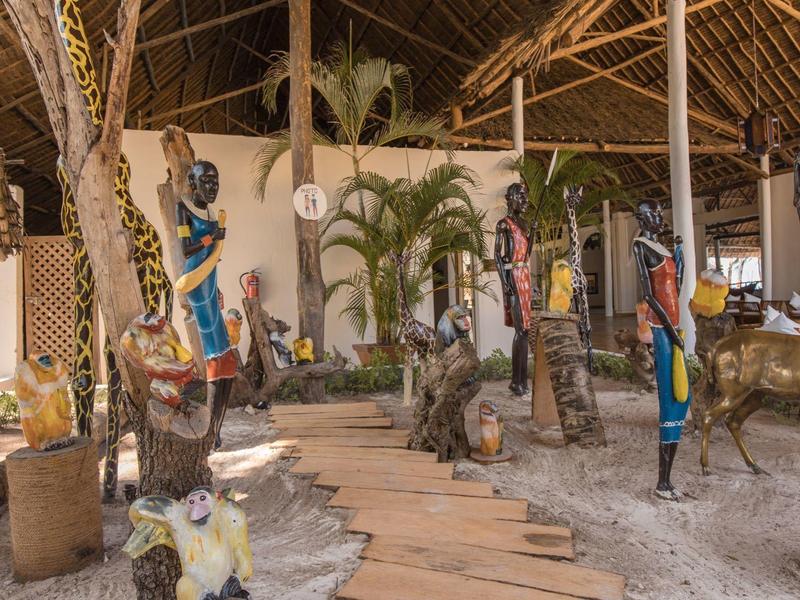 Überdachter Weg mit Holzplanken und afrikanischer Kunst in tropischer Umgebung.