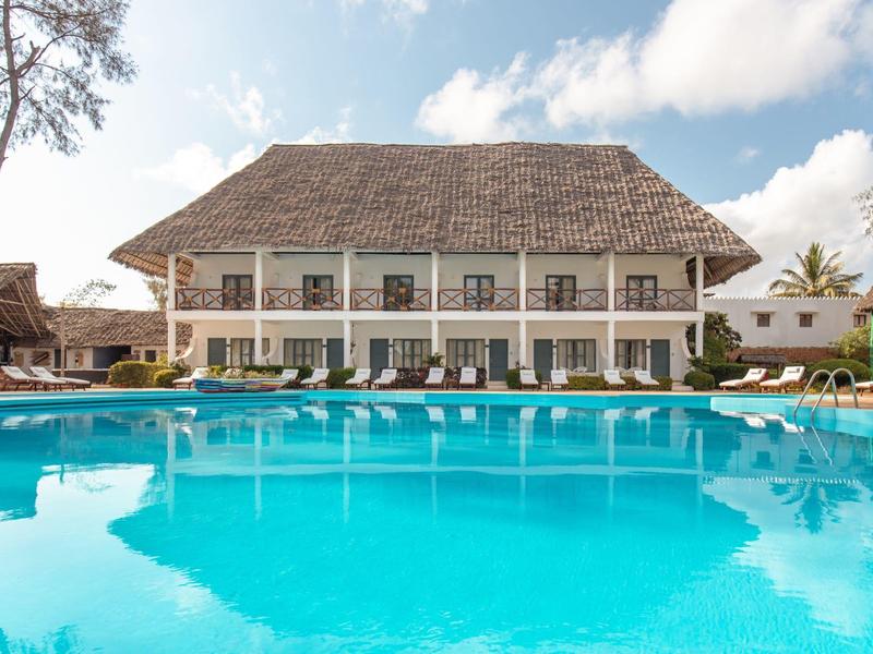 Hotel mit zwei Stockwerken und Balkon, umgeben von einem großen blauen Pool bei sonnigem Wetter.