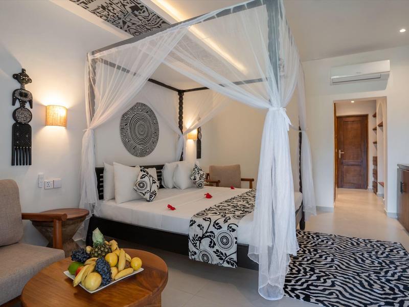 Helles Hotelzimmer mit Baldachinbett, dekorativen Kissen und Früchtekorb auf Holztisch.