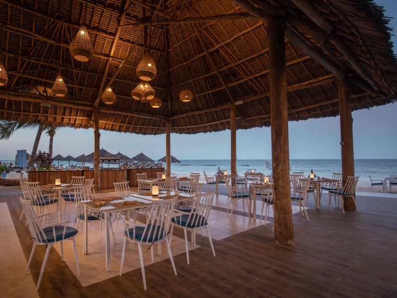 Offener Strandrestaurantbereich unter einem Strohdach mit weißen Stühlen und Blick auf das Meer.