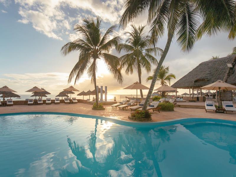 Pool mit Sonnenliegen und Palmen am Strand bei Sonnenuntergang.