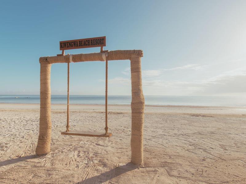 Eine einsame Schaukel auf einem weiten Sandstrand mit klarem Himmel und ruhigem Meer im Hintergrund.