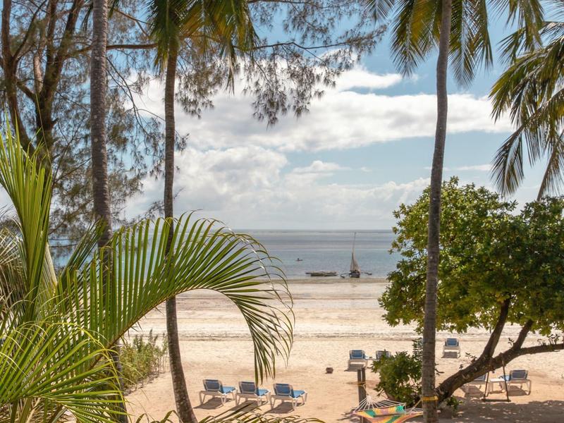 Strandansicht mit Palmen, Liegestühlen und ruhigem Meer im Hintergrund bei sonnigem Wetter.