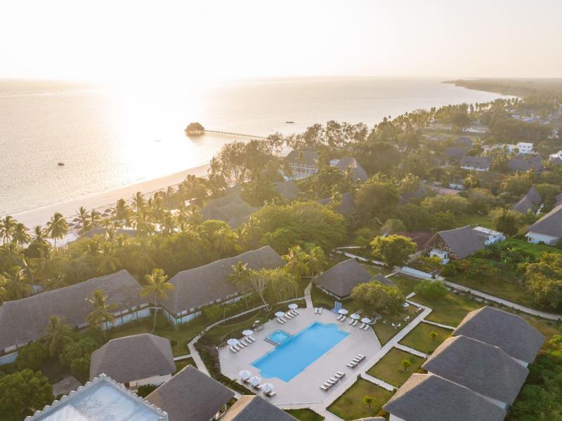 Luftaufnahme eines Resorts mit Pool, umgeben von Gebäuden und Blick auf das Meer bei Sonnenuntergang.