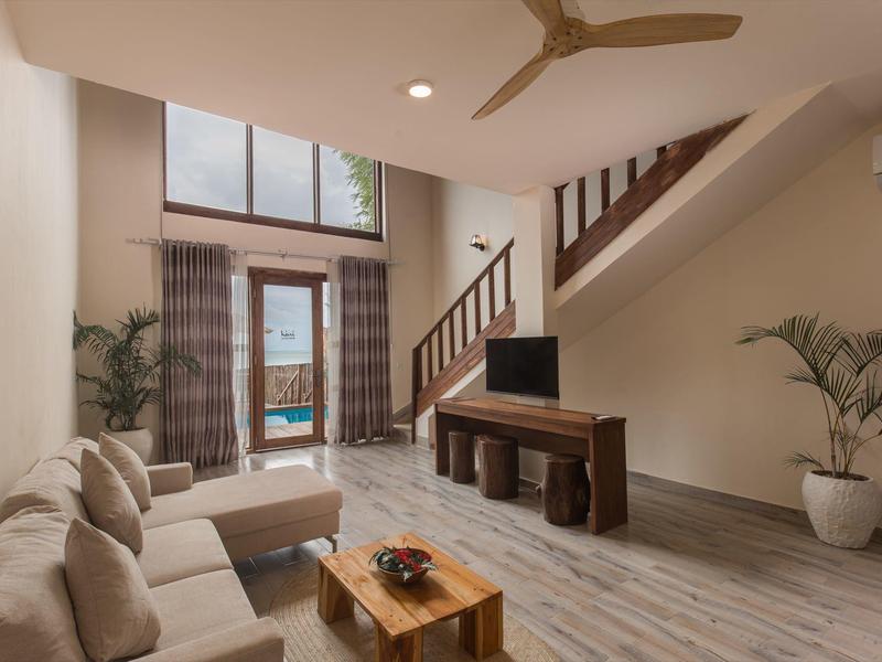 Wohnzimmer mit beiger Couch, Holztisch, TV und Treppe zu einer oberen Etage mit Blick auf den Pool.