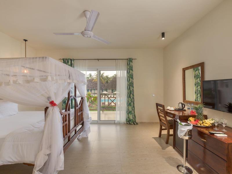 Helles Hotelzimmer mit Doppelbett, Deckenventilator, Schreibtisch und Zugang zu Balkon mit Gartenblick.