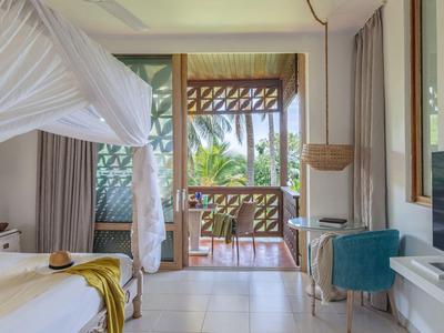 Helles Hotelzimmer mit Doppelbett, Balkon mit Sicht auf tropische Pflanzen und Holzterrasse.