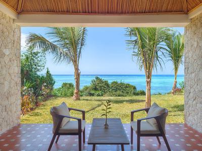 Blick von einer überdachten Terrasse mit Stühlen auf tropischen Garten und Meer.