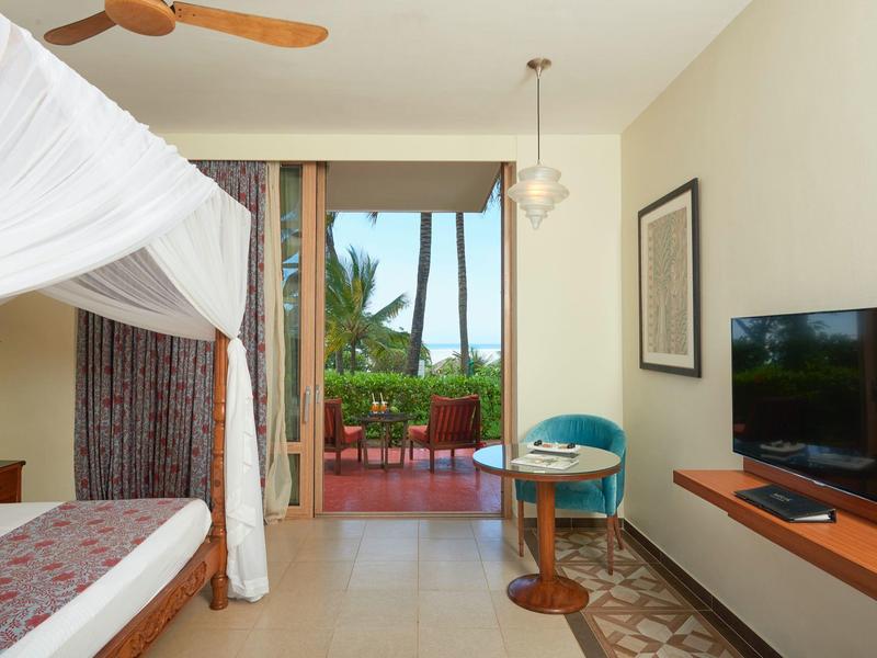 Helles Schlafzimmer mit großem Bett, TV, Balkon und Blick auf Palmen und Terrasse.