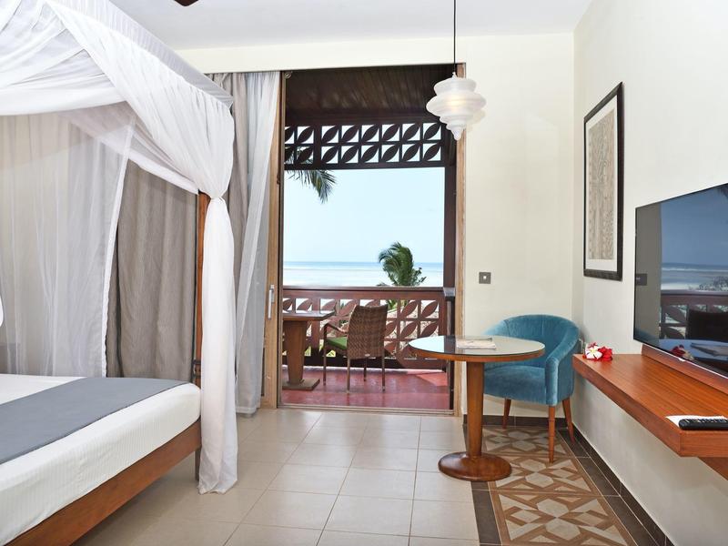 Helles Hotelzimmer mit Himmelbett, TV, kleinem Tisch, Stuhl und Balkon mit Meerblick.