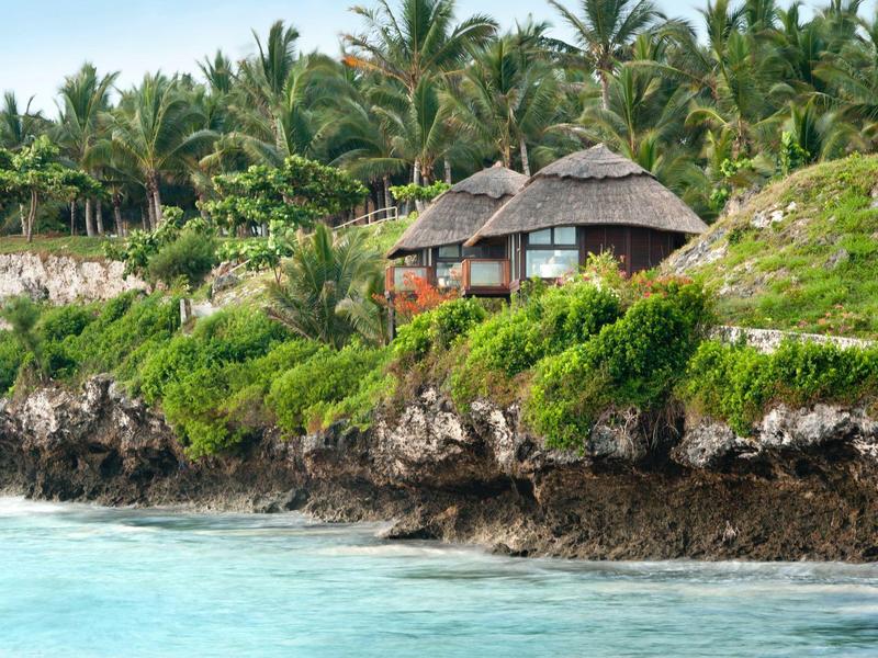 Kleine strohgedeckte Hütten auf felsiger Küste mit grünem Buschwerk und Palmen im Hintergrund.