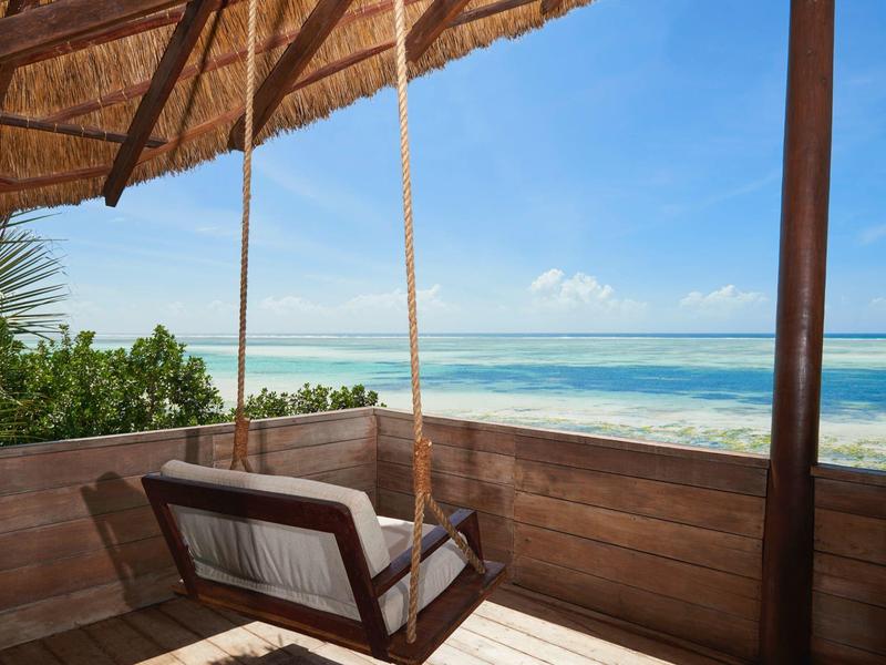 Holzterrasse mit Hängesessel und Blick auf ruhiges, türkisfarbenes Meer unter blauem Himmel.