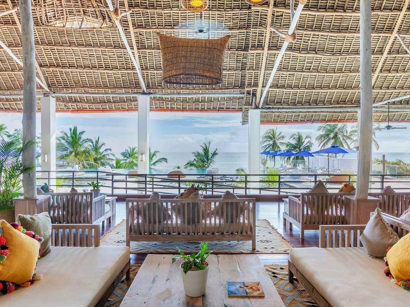 Offener Strandlounge-Bereich mit Holzmöbeln, bunten Kissen und Palmen im Hintergrund.