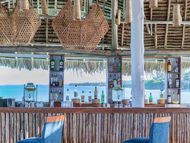 Helle Bar mit Holztheke, blauen Hockern und großen geflochtenen Lampenschirmen unter Strohdach.