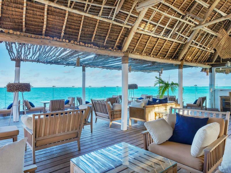 Offener Strandpavillon mit Holzmöbeln, Strohdach und Blick aufs blaue Meer.