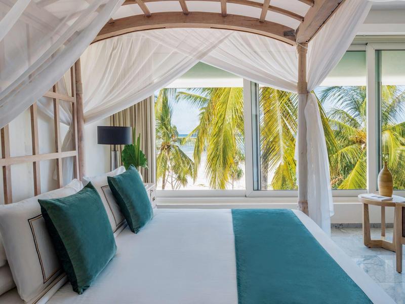 Helles Schlafzimmer mit großem Bett, weißen Vorhängen und Blick auf grüne Palmen durch Fenster.