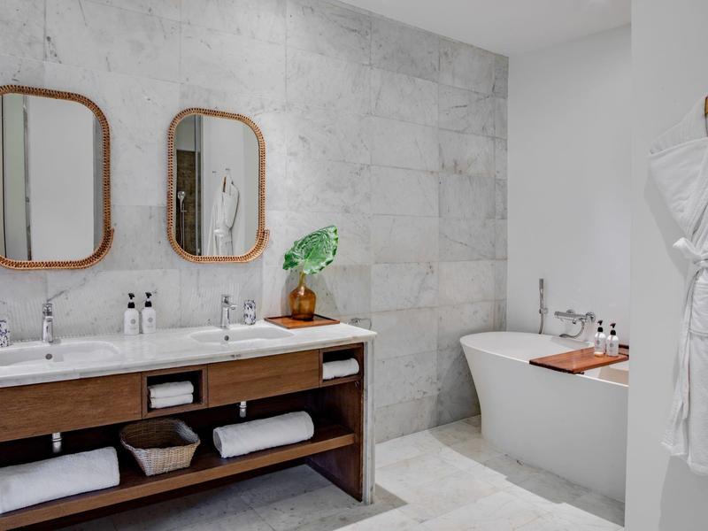 Modernes Badezimmer mit grauen Fliesen, Doppelwaschbecken, Spiegeln, freistehender Badewanne und Bademantel.