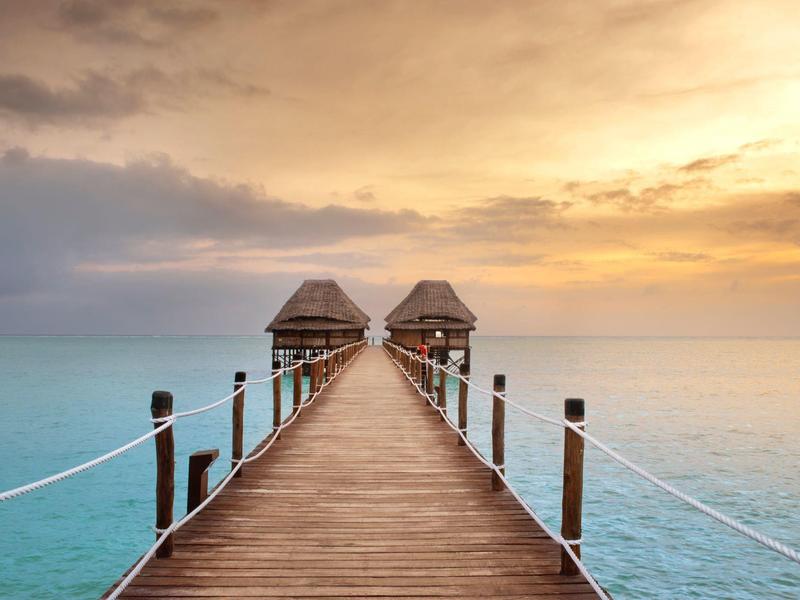 Holzsteg mit zwei Bungalows am Ende, ruhiges blaues Meer, goldener Himmel bei Sonnenuntergang.