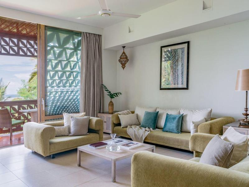 Wohnzimmer mit beigen Sofas, blauen Kissen, Holzlampe und Pflanzen vor Balkon mit grünen Gittern.